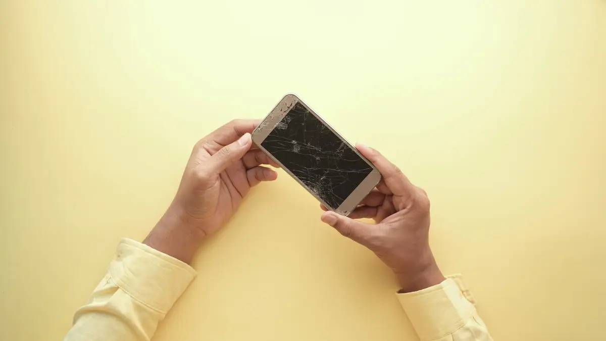 Close-up of hands holding a cracked smartphone against a yellow backdrop, highlighting mobile technology issues.