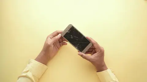 Close-up of hands holding a cracked smartphone against a yellow backdrop, highlighting mobile technology issues.
