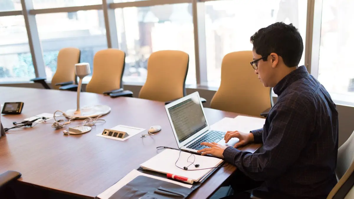 A professional adult sitting at a desk with a laptop in a bright, modern conference room.