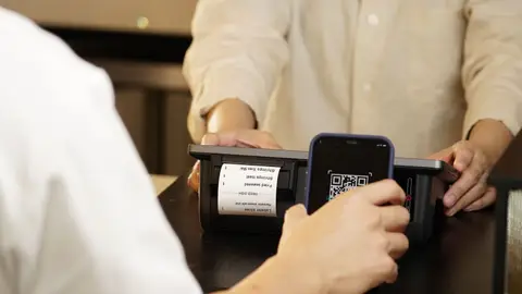 Two individuals engaged in a contactless payment using a QR code scanner at a retail counter.