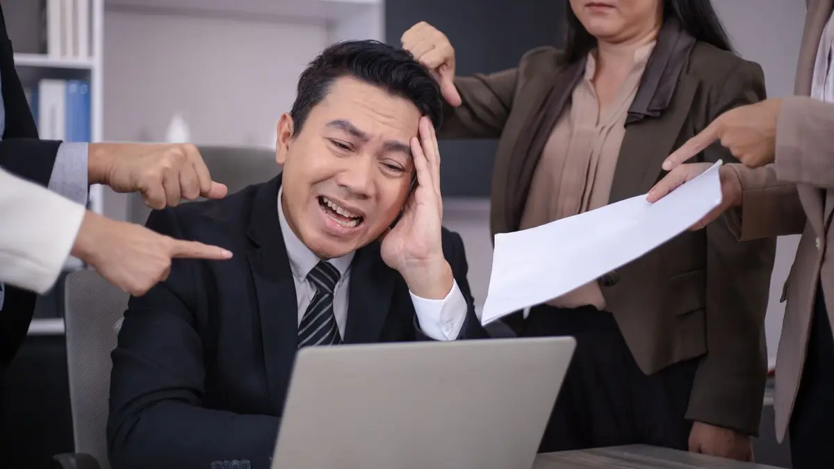 An Asian businessman looks stressed as colleagues point fingers and give him paperwork in a modern office.