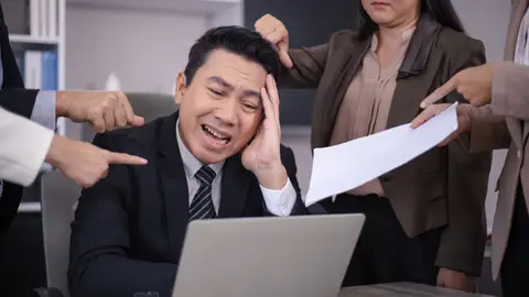 An Asian businessman looks stressed as colleagues point fingers and give him paperwork in a modern office.