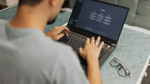Man using laptop with AI interface, typing with focused attention. Indoors with eyewear beside.