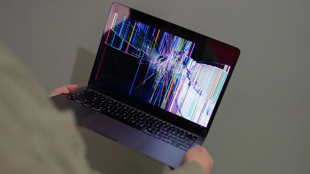A broken laptop screen displayed with colorful glitch being held by a person.