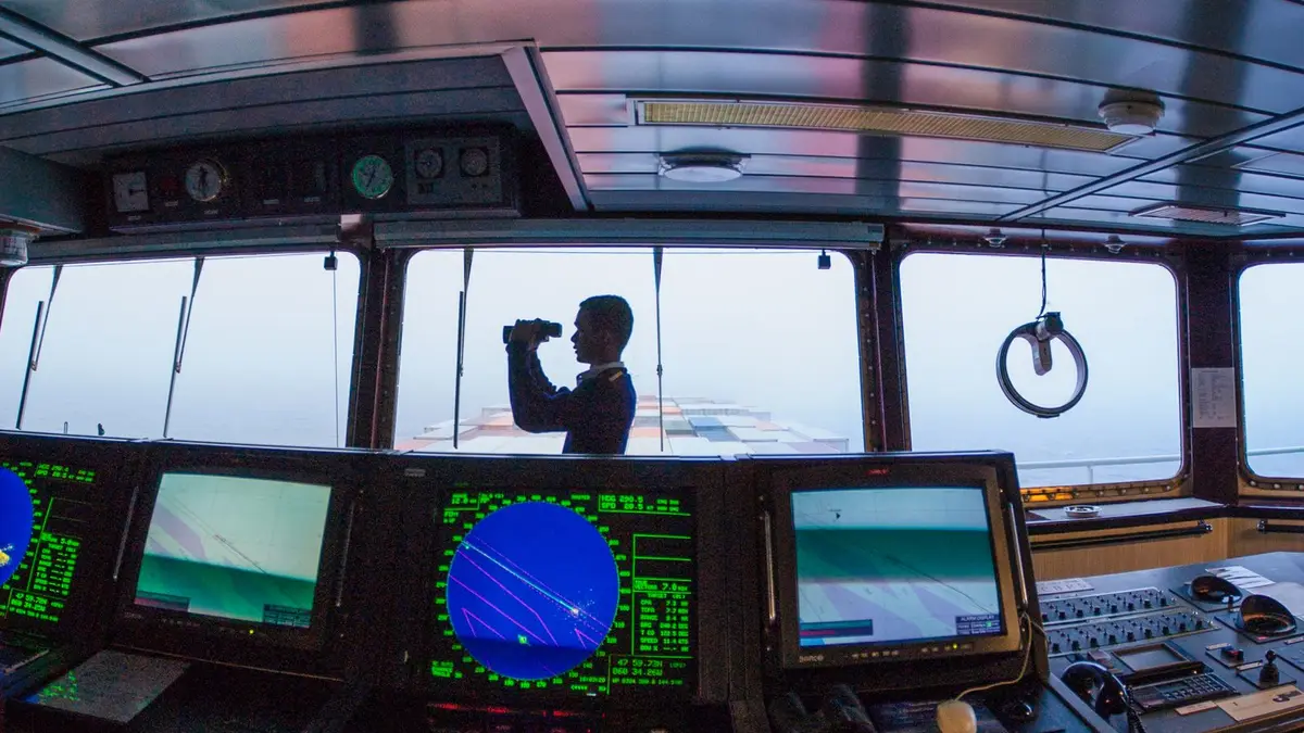 Silhouette of a navigator using binoculars on a cargo ship's bridge, surrounded by navigation equipment.