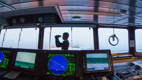 Silhouette of a navigator using binoculars on a cargo ship's bridge, surrounded by navigation equipment.