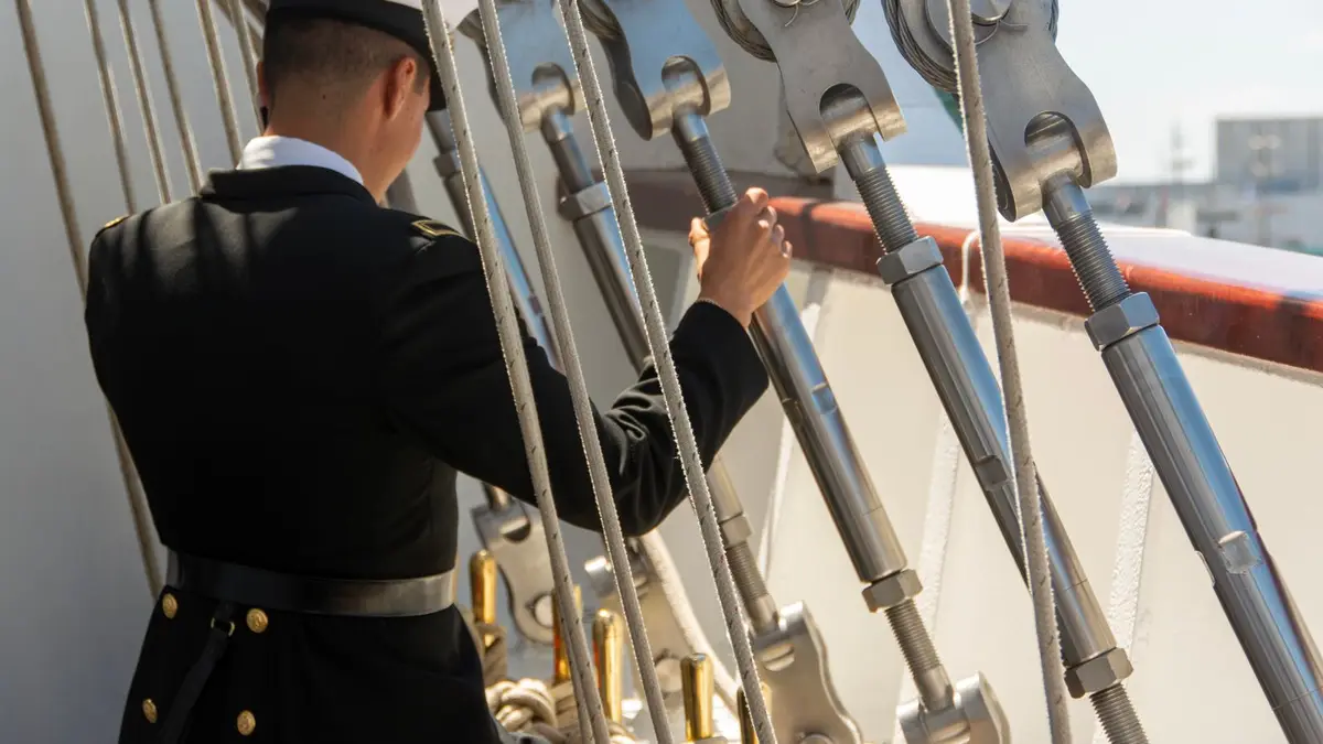 A sailor in uniform adjusts ropes on a ship deck during the day.