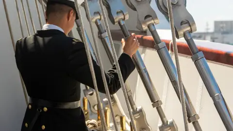 A sailor in uniform adjusts ropes on a ship deck during the day.