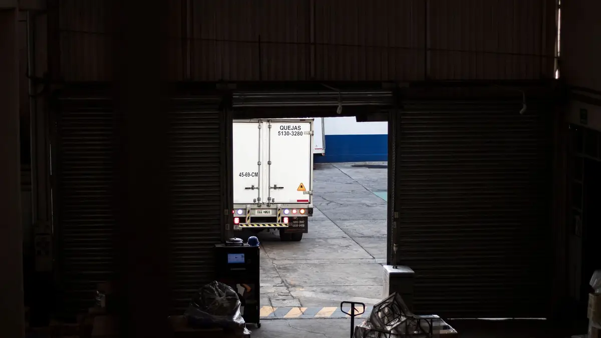 A white delivery truck exits a dimly lit warehouse loading dock, surrounded by packages and pallets.