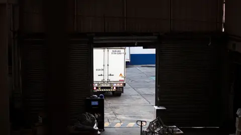 A white delivery truck exits a dimly lit warehouse loading dock, surrounded by packages and pallets.