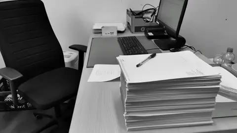 Black and white image of a cluttered office desk with a computer, chair, and paperwork.
