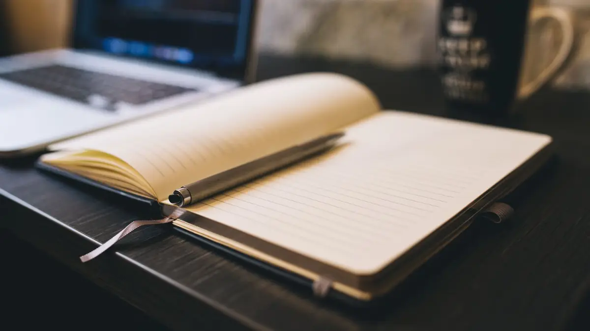Open notebook with a pen on a wooden desk beside a laptop, ideal for blogging and work-related themes.