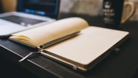 Open notebook with a pen on a wooden desk beside a laptop, ideal for blogging and work-related themes.