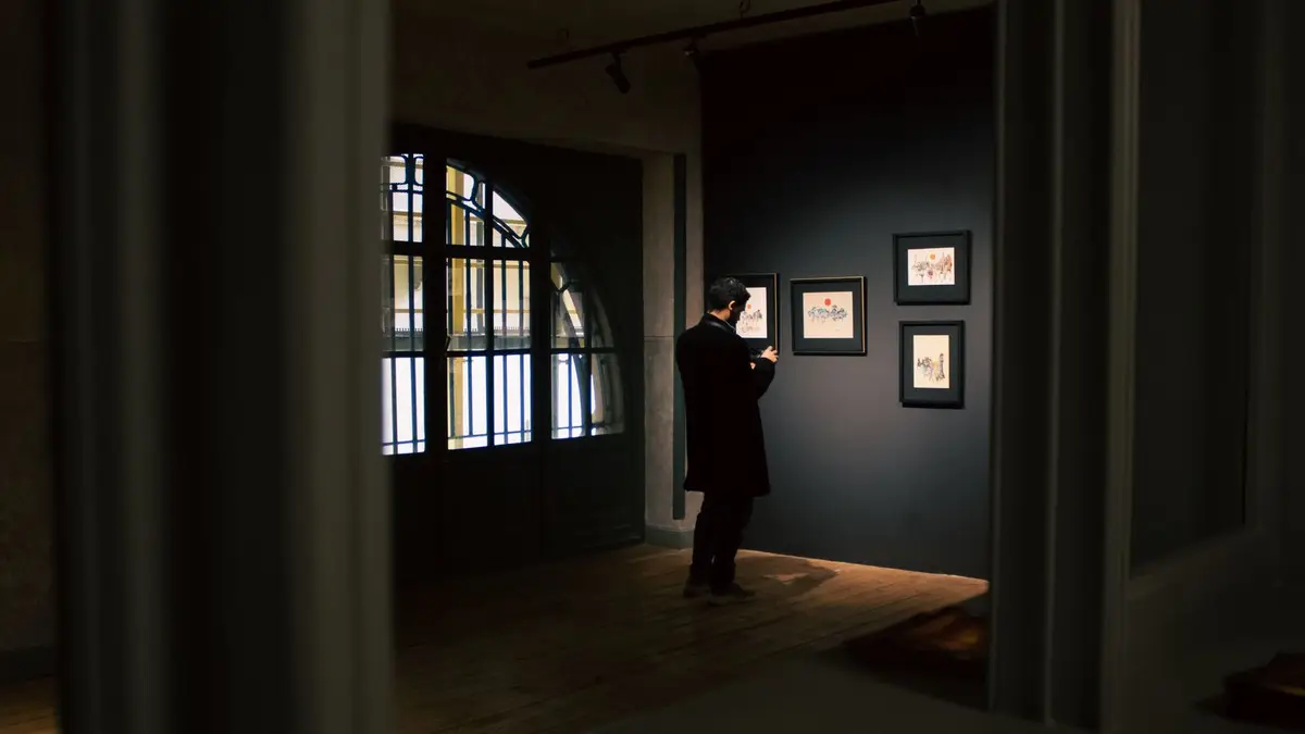 A man in a gallery analyzes framed art in dim lighting, creating a sophisticated atmosphere.