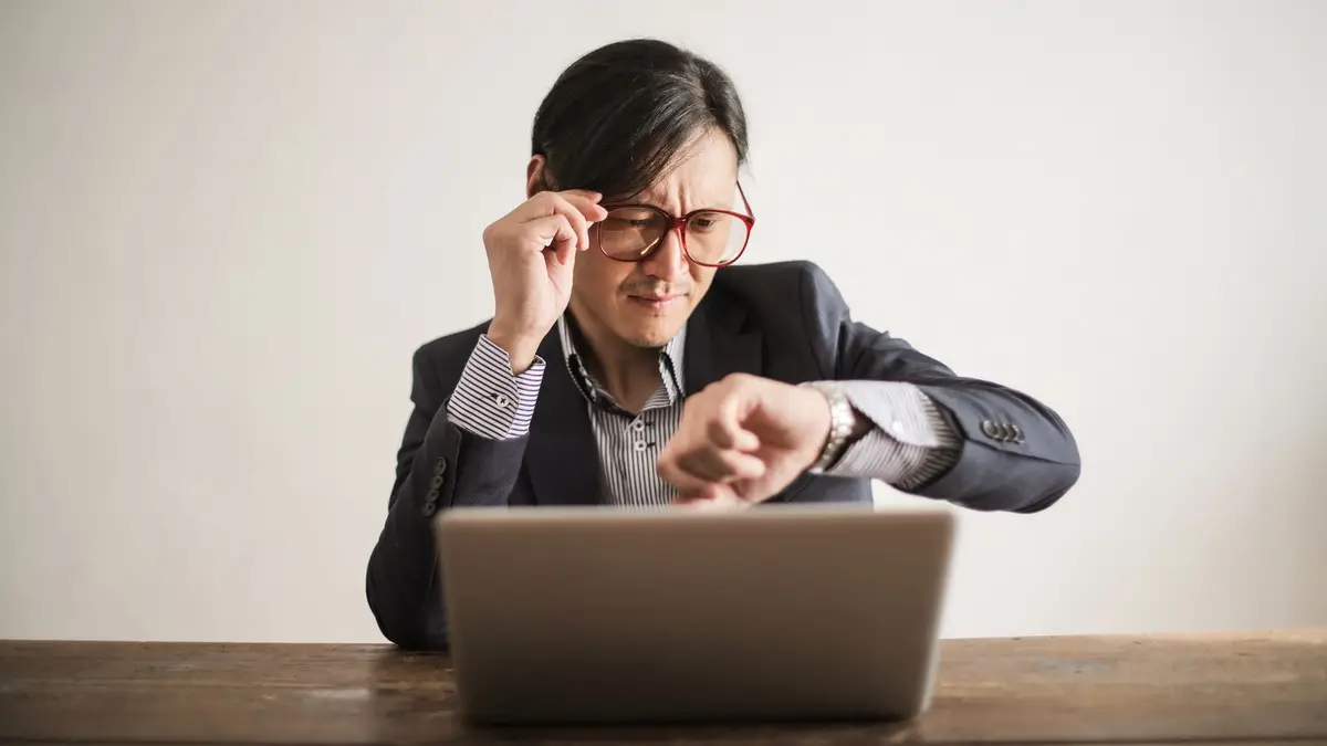 Young frowning man in suit and glasses looking at wristwatch while waiting for appointment sitting at desk with laptop