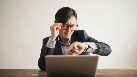 Young frowning man in suit and glasses looking at wristwatch while waiting for appointment sitting at desk with laptop