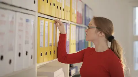 A focused woman in the office organizing and searching through colorful binders.