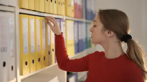 Side view of contemplating female assistant in casual style standing near shelves and choosing file with documents