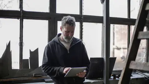 Serious adult bearded workman wearing workwear standing near window and browsing tablet while working in workshop