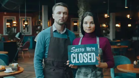 Two café owners holding a 'Sorry We're Closed' sign inside their restaurant.