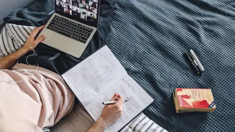 Adult engaged in online class with a laptop and notebook while sitting in a cozy bed setting.