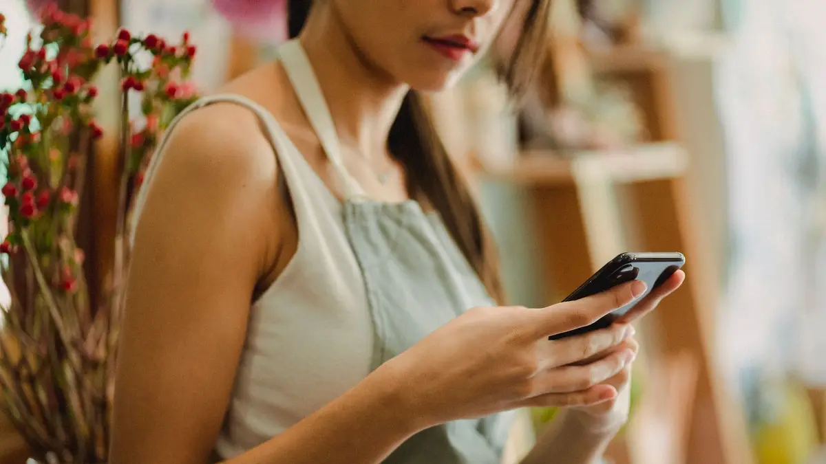 Crop anonymous florist in apron browsing mobile phone while working in cozy floral shop