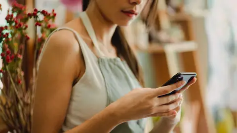 Crop anonymous florist in apron browsing mobile phone while working in cozy floral shop