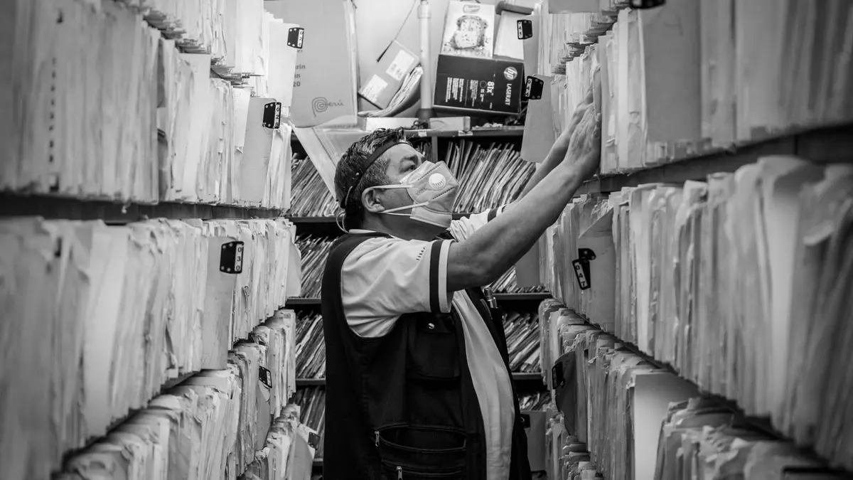 Elderly man sorts through files in an office aisle wearing a face mask.