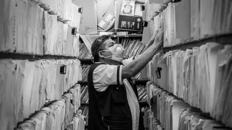 Elderly man sorts through files in an office aisle wearing a face mask.