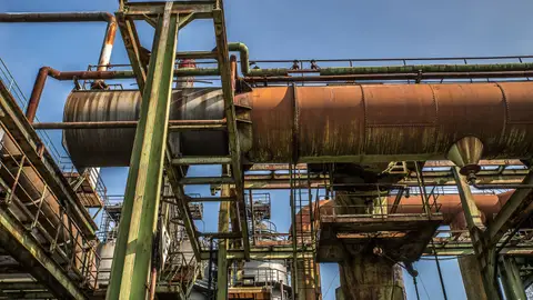 A low angle view of a rusty old industrial structure with pipes and beams against a clear blue sky.