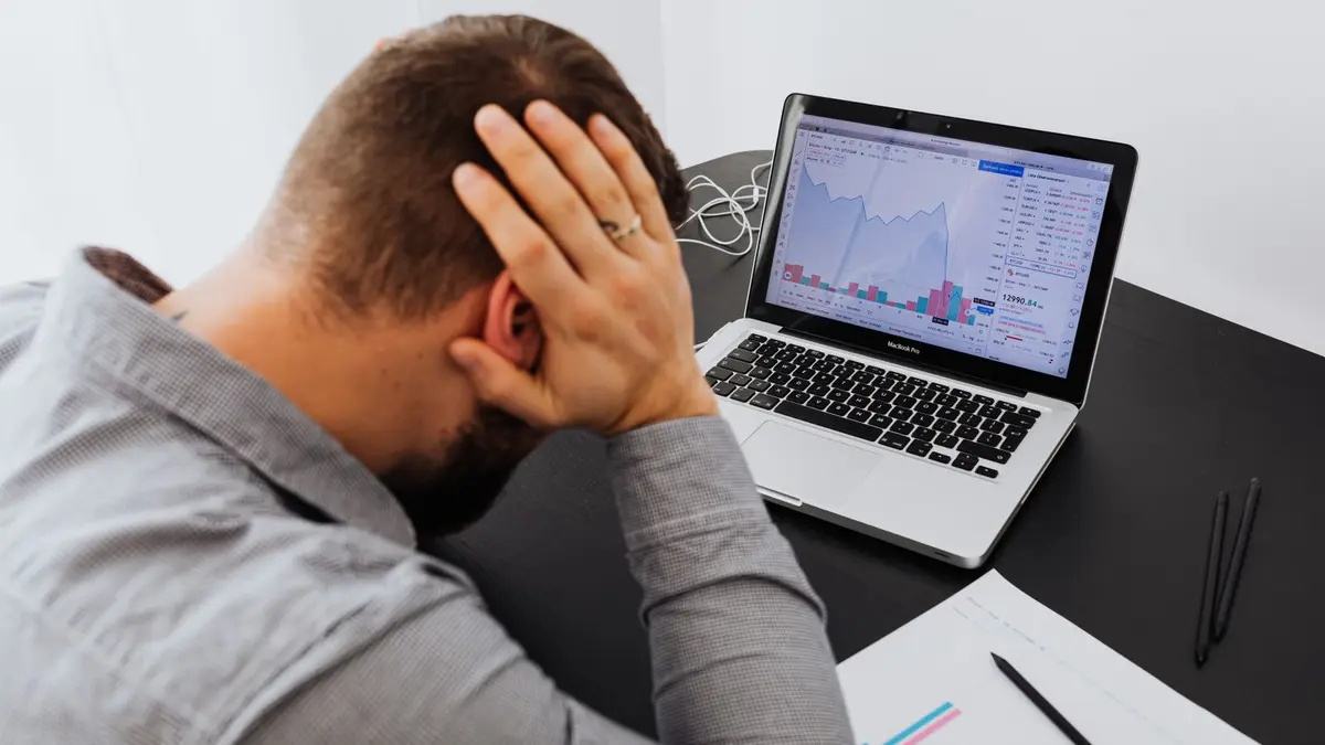 Stressed man at desk looking at declining stock charts on laptop, indicating financial loss.