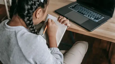 Back view of little schoolgirl writing in notepad while studying at desk with laptop