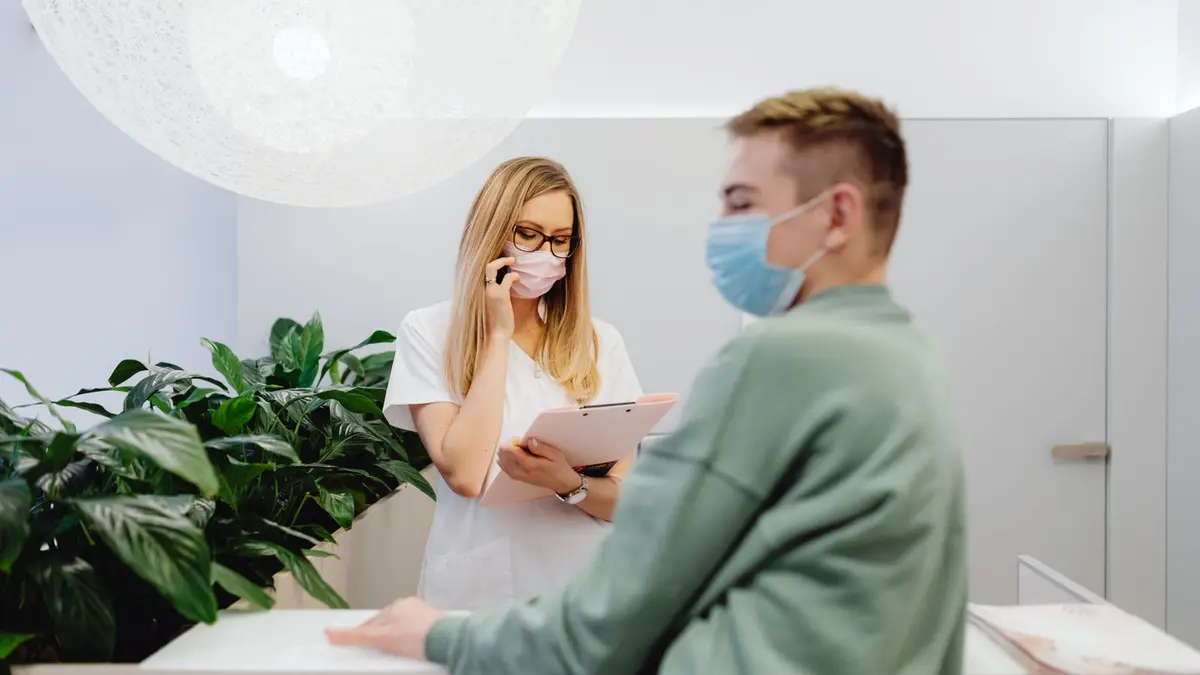 Healthcare worker and patient wearing masks at clinic front desk during pandemic, highlighting safety measures.