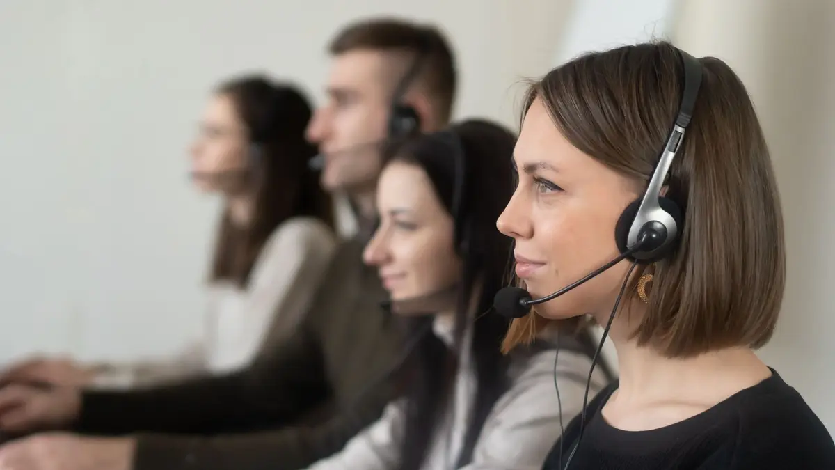 Side view of young coworkers in casual clothes sitting in light workplace with headphones with microphone while surfing on laptop