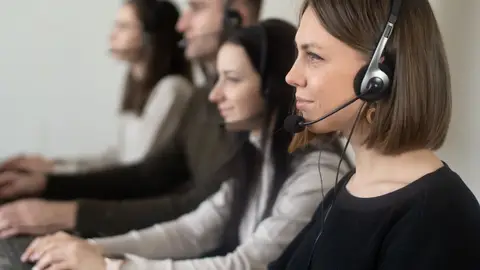 Side view of young coworkers in casual clothes sitting in light workplace with headphones with microphone while surfing on laptop