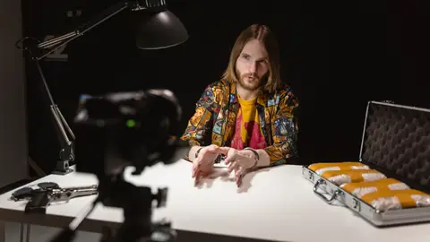 A dramatic scene depicting a man in an interrogation room discussing on camera, with a suitcase and gun visible.