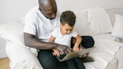 A father sits with his young son on a sofa, teaching him how to use a laptop in a cozy home setting.
