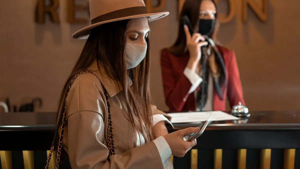 Woman wearing a mask checking her phone at a hotel reception during COVID-19.