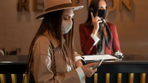 Woman wearing a mask checking her phone at a hotel reception during COVID-19.
