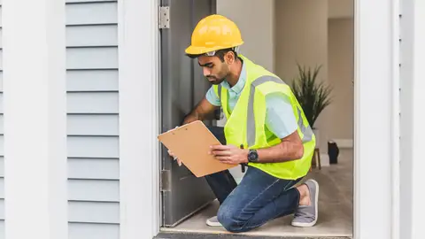 A home inspector wearing safety gear examines a house interior for safety compliance.