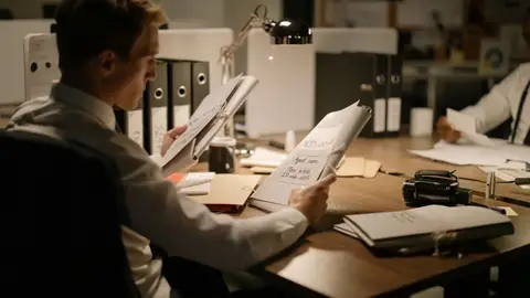 Two detectives reviewing documents in a dimly lit office setting, focused and professional.