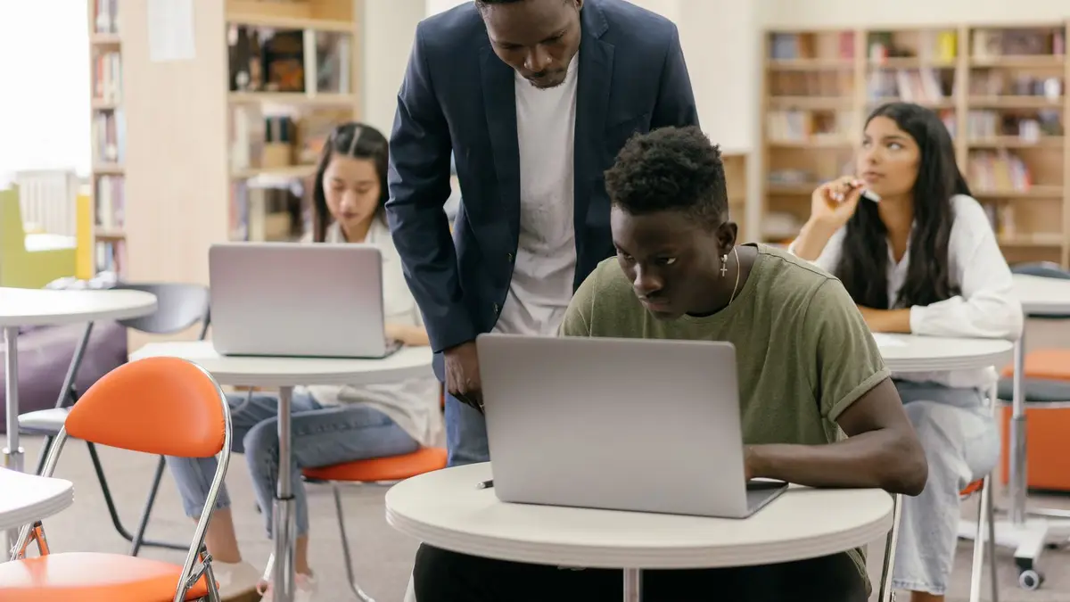 Students learning in a classroom setting with a teacher assisting and laptops on desks, creating an interactive education environment.