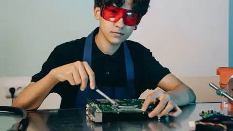 Young man in apron and goggles working on a motherboard indoors.