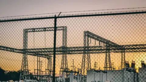 Industrial electric power substation photographed at sunset with fence in the foreground.